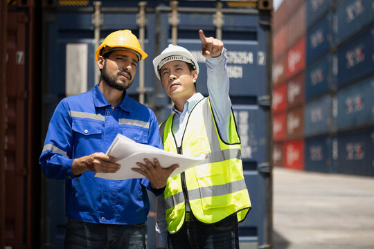Logistics engineer and supervisor reviewing shipment documents at container yard, coordinating global cargo and supply chain operations with safety and teamwork in industrial port environment.