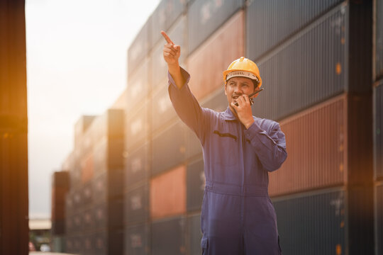 Logistics worker using walkie talkie and pointing in container yard, coordinating cargo and shipping at industrial port, concept of teamwork, safety, transportation and global supply chain.