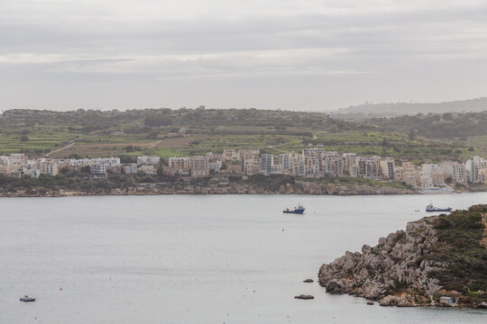 View of the tranquil bay waters reflecting the somber sky, embracing the coastal buildings nestled against the distant green hills, Il-Mellieha, Malta.