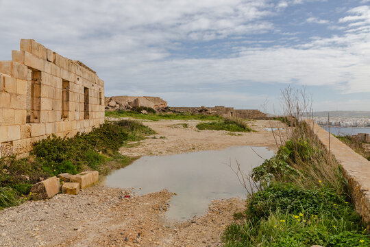 View of aged stone walls meet the horizon under a vast sky, reflected in a still puddle, contrasting textures and natural hues, Il-Mellieha, Malta.