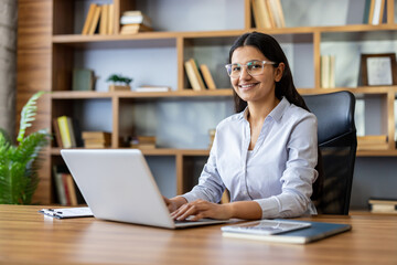 Young indian woman wearing eyeglasses smiling at the camera, working on a laptop at a wooden desk in a modern office with bookshelves in the background © Liubomir