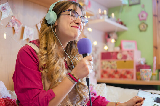 A young woman with glasses and headphones sings into a microphone while looking at a laptop in her decorated room.