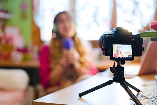 A young woman wearing headphones speaks into a microphone while recording a video with a camera on a tripod.