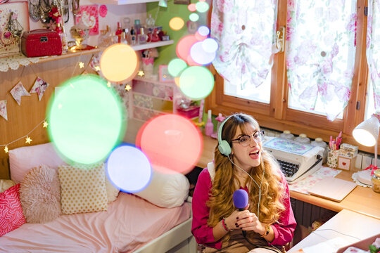 A young woman with headphones sings into a microphone in her decorated bedroom, surrounded by colorful bokeh lights.
