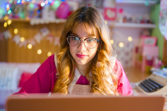 A young woman with glasses and blonde hair looks intently at a laptop screen in a room decorated with colorful lights.