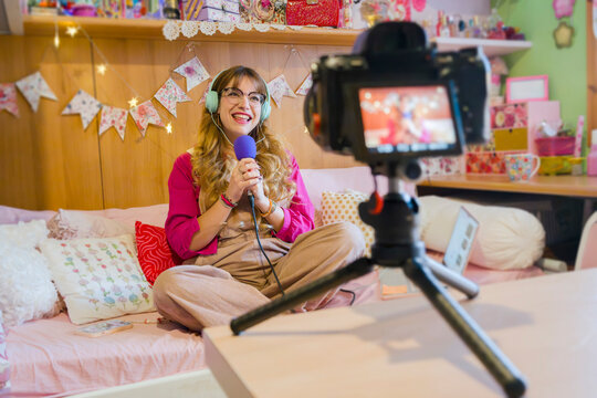 A young woman with headphones and glasses records herself speaking into a microphone, sitting on a bed in a decorated room.