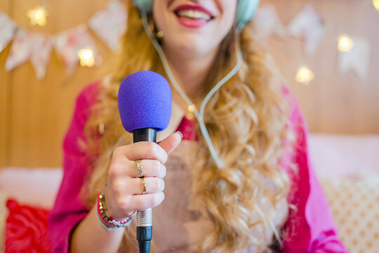 A young woman with blonde hair and headphones holds a microphone, smiling as she records audio.