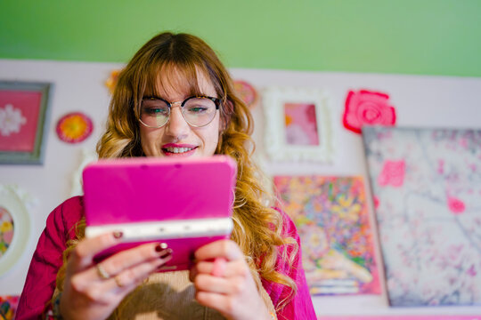 A young woman with glasses smiles as she plays a handheld gaming device in a room decorated with colorful artwork.