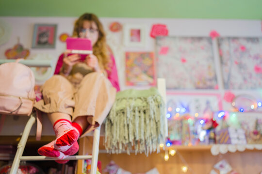 A young woman sits on a ladder, engrossed in her handheld electronic device, surrounded by a whimsical, decorated room.