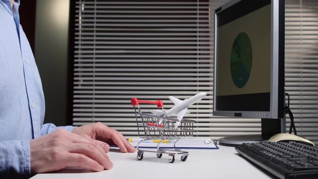 Person's hand holding a mini shopping cart with a toy airplane while sitting at a desk with a computer displaying a pie chart, symbolizing online travel booking and e commerce