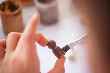 Close-up of a hand applying paint to a small wooden figurine with a brush, surrounded by jars of...