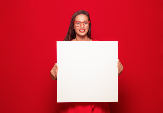 Promoting women holding white board, isolated on red background. Amused girl showing blank empty paper billboard with blank space for text.