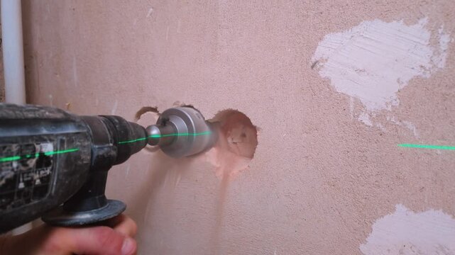 Professional worker using a powerful hammer drill with a core bit to make a hole in a concrete wall for an electrical outlet, guided by a green laser level for precise alignment