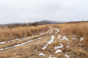 Fototapeta premium Snow-covered dirt path winding through dry grassland with distant hills under overcast sky in a rural landscape during winter season