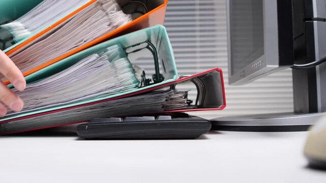 Stacked office binders overflowing with paperwork creating a feeling of being overwhelmed and signifying a heavy workload on an office desk next to a computer monitor