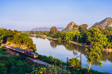 Train Journey along River Kwai. Scenic view of train traversing famous historic curve of Railway line surrounded by green hills at Tham Khao Pun Temple viewpoint. World War II Heritage Rail, Thailand. © JinnaritT