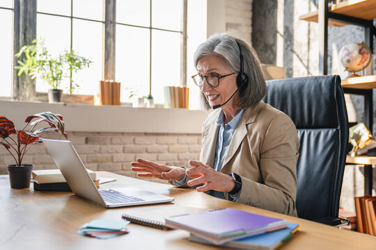 Virtual communication. Smiling mature woman remote working interpreter tech support consultant in headset with mic talk to client via video call conference app on pc engaged in online event.