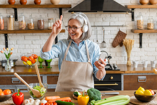Happy mature woman dance and cook in the kitchen, listen popular track, having fun, moving to favorite music while prepare healthy dieting vegetarian salad at home. Chore, lifestyle, weekend activity