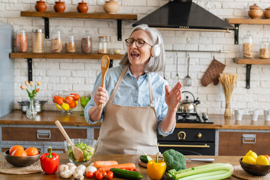 Excited mature woman singing and dancing in modern kitchen at home, happy woman holding spatula as microphone, dancing, listening to music, having fun with kitchenware, preparing breakfast