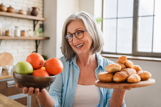 Good nutrition for weight loss concept. Happy mature senior lady holding plates with fruits and desserts at home, making choice between healthy and unhealthy foods, choosing balanced diet