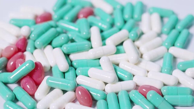 Assortment of green, white, and pink pharmaceutical capsules spins in a seamless loop. This macro footage on white surface visualizes medical treatment, drug manufacturing, and clinical research.