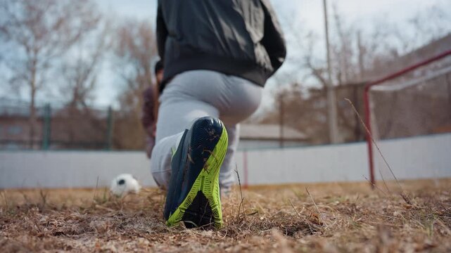 close-up cleats striking ball into goal, white male striker drives powerful shot on muddy urban pitch with worn net and autumn light, intense footwork and focused stance capture raw athletic grit