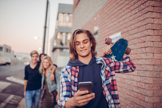 Young man with skateboard using phone on city street