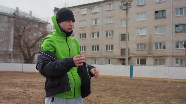 white male adjusts jacket and focuses on pregame routine on empty urban pitch, hands fidgeting with zipper, calm breath, mental preparation before training, muted sky and apartment backdrop, steady