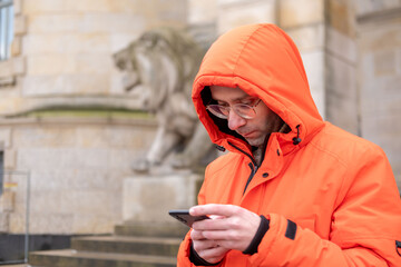 young Man in bright orange winter jacket and glasses using smartphone outdoors, male character in hooded parka texting on mobile phone, urban background with historical building, digital lifestyle © kittyfly