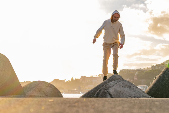Energetic young man in white hoodie walking on concrete breakwaters against blue sunset sky, embodying youth, freedom, positive aspiration, capturing freedom, travel, quiet awe nature&rsquo;s grandeur