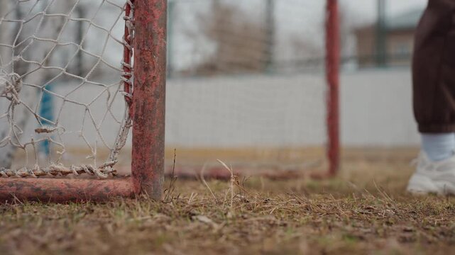 foot striking near rusty goalpost on brown field, caucasian player sneakers and white socks close to net, low camera angle captures impact, follow-through and gritty urban pitch details