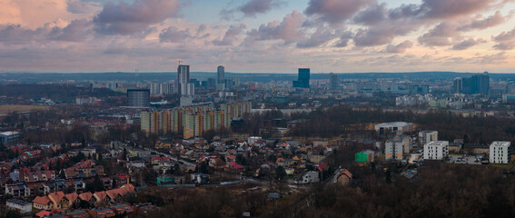 Aerial view of Katowice, Poland, shows suburban houses, panel blocks, and modern high rises. Altus Tower and .KTW cluster stand near cranes under a pastel sunset sky.