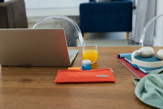 Dining table workspace showing laptop, glass of orange juice, orange pouch, notebooks, headphones