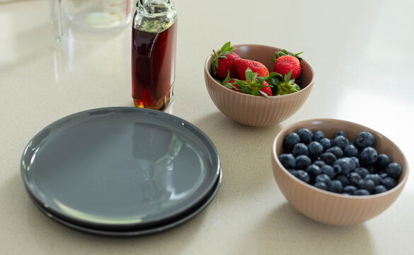 Two stacked gray plates sitting beside glass syrup bottle and beige bowls of berries on countertop
