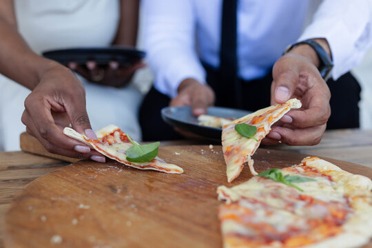 Hands are grabbing thin-crust pizza from wooden board on table, cheese crumbs are falling