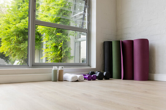 Rolled mats, foam roller and dumbbells are resting by open window, towels and bottles nearby