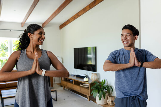 African couple practicing paired yoga in living room by TV, wearing athletic wear and smartwatches