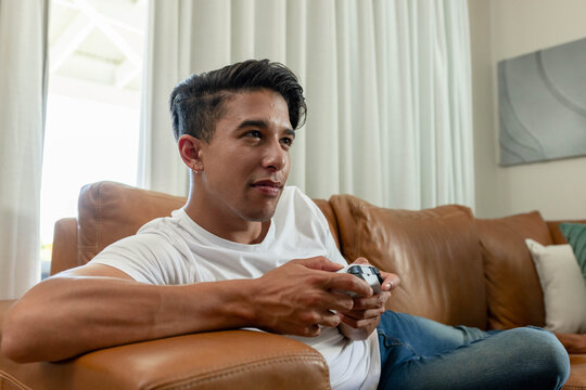 African American man reclining on sofa at home holding wireless controller in white tee and jeans