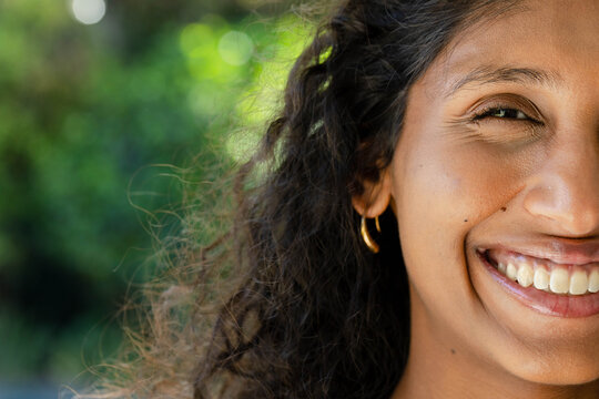 African American woman smiling broadly, showing teeth with curly hair, gold hoop and bokeh in park