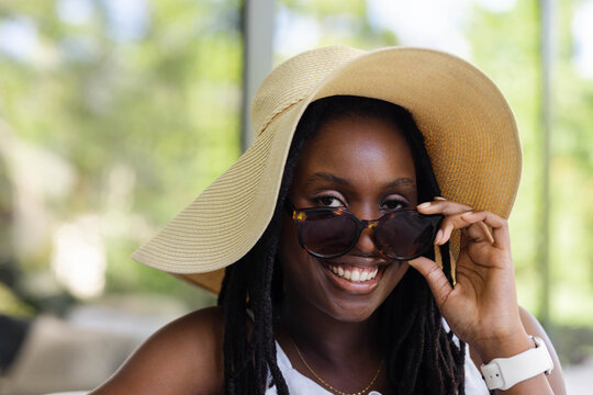 Woman wearing straw sun hat lowering tortoiseshell sunglasses while smiling in bright sunroom