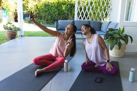 Diverse female friends taking phone selfie on yoga mats on tiled patio in activewear and watches
