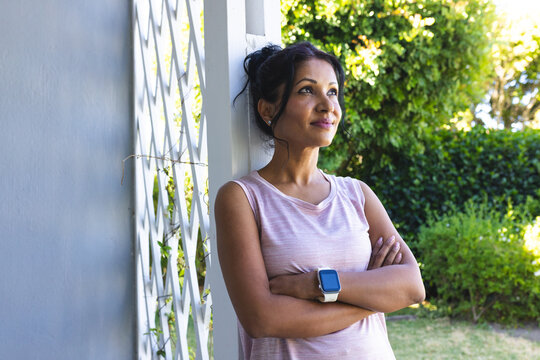Indian woman leaning against white lattice trellis on porch, wearing light pink top and smartwatch
