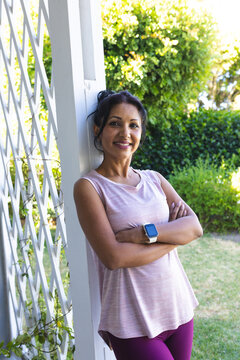 Woman leaning on white trellis post in porch, in pink top and purple leggings, checking smartwatch