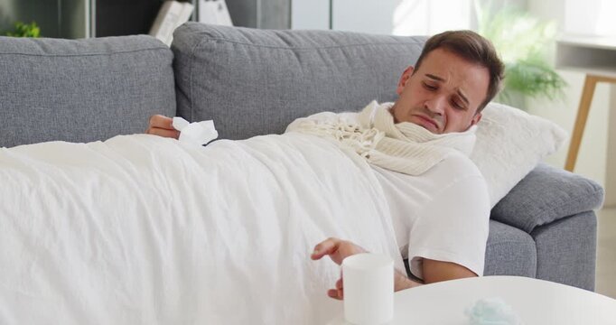 Sick man with virus cold blowing nose. Flu symptoms with fever, sneeze and cough, using tissue while resting under blanket with scarf at home. Useful for healthcare and seasonal illness.