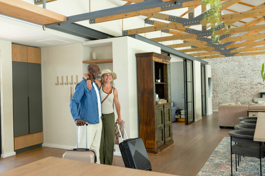 Mature couple standing near loft entrance, holding rolling suitcases, wearing hat and denim shirt