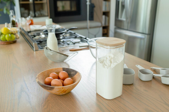 Flour canister is sitting on wooden island in home kitchen, with eggs, milk bottle, sieve