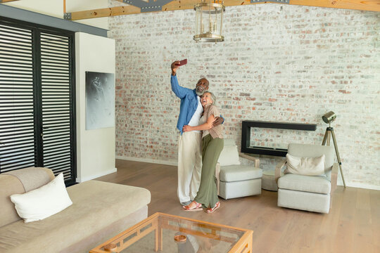 Couple hugging and taking selfie on smartphone in living room, wearing blue shirt and olive pants