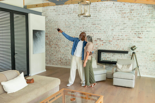 Couple with senior man wearing denim jacket and woman posing selfie in living room with smartphone