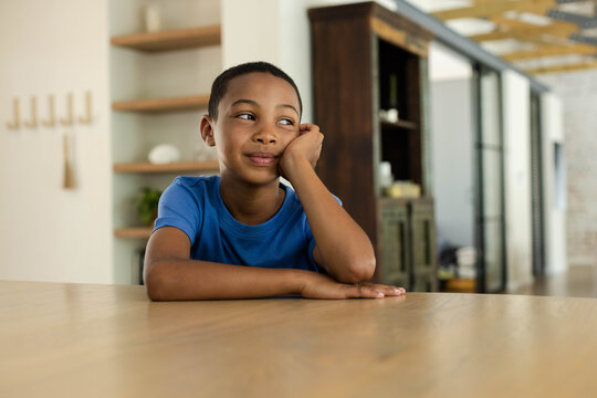 African American schoolboy sitting at wooden table at home, resting head, wearing blue T-shirt