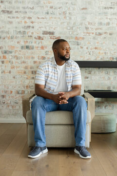 Adult African American man sitting on armchair in studio, wearing shirt, jeans, looking right